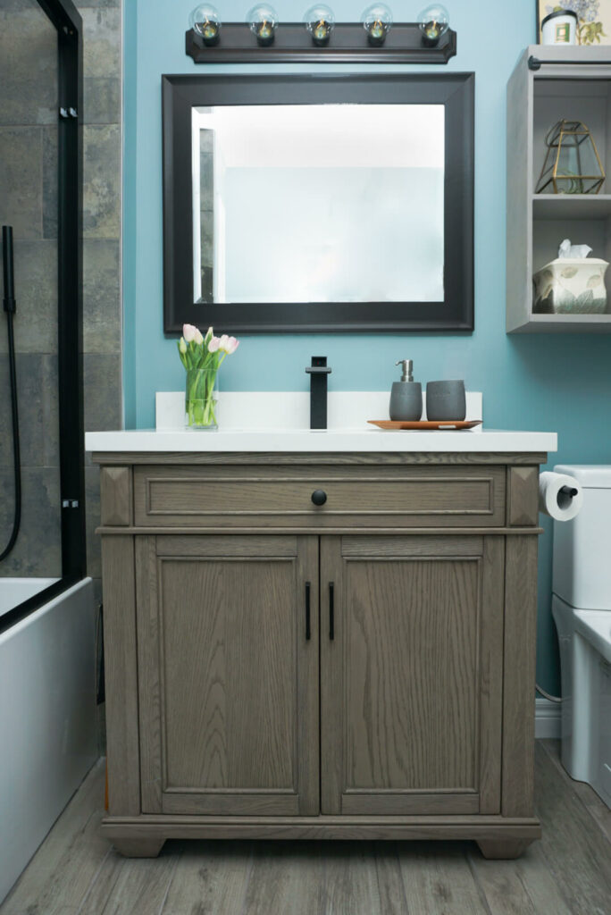 Modern single vanity with wooden cabinets, white counter, and black sink. A single square, black-framed mirror hangs above against a teal wall.