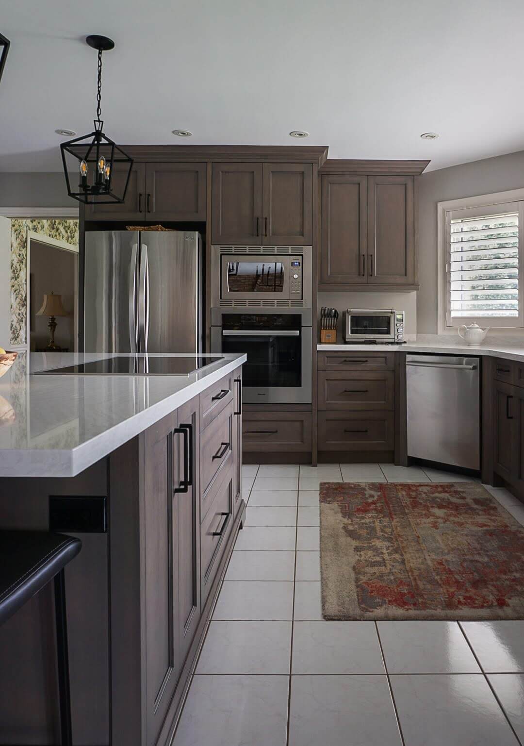 Sideview of a renovated kitchen with white marble island countertop, silver appliances, and brown wooden cabinets and decorative rug